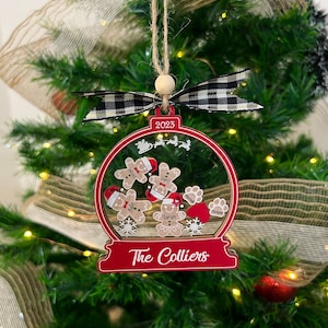 May include: A red and clear snow globe ornament featuring the text 2023 and The Colliers. Inside are gingerbread figures with names, paw prints, a red heart, and white snowflakes. It is topped with a black and white gingham bow and twine.