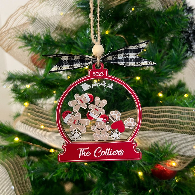 May include: A red and clear snow globe ornament featuring the text 2023 and The Colliers. Inside are gingerbread figures with names, paw prints, a red heart, and white snowflakes. It is topped with a black and white gingham bow and twine.