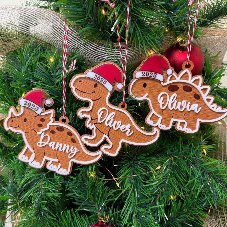 Op de afbeelding: Drie houten dinosaurusornamenten met een bruine afwerking, elk met een rood-witte kerstmuts waarop het jaartal 2025 staat gedrukt. De ornamenten zijn gepersonaliseerd met de in witte letters geschreven namen Danny, Oliver en Olivia aan rood-wit koord.