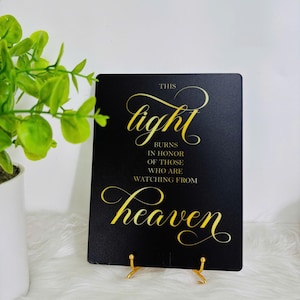 May include: A black rectangular sign with gold script that reads "This light burns in honor of those who are watching from heaven." The sign is displayed on a gold metal stand, with a green plant in a white pot in the background.