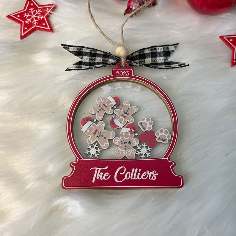May include: A red and clear snow globe Christmas ornament featuring the text 2023 and The Colliers. Inside are wooden gingerbread figures with names, paw prints with names, a red heart, and white snowflakes, topped with a black and white plaid bow.