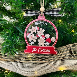 May include: A red and clear snow globe ornament featuring the text 2023 and The Colliers. Inside are gingerbread figures with names, paw prints with names, a red heart, and snowflakes. It is topped with a black and white buffalo plaid ribbon.