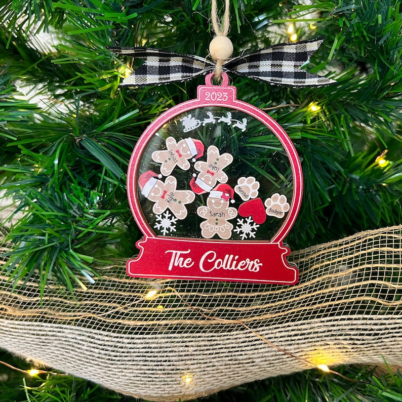 May include: A red and clear snow globe ornament featuring the text 2023 and The Colliers. Inside are gingerbread figures with names, paw prints with names, a red heart, and snowflakes. It is topped with a black and white buffalo plaid ribbon.