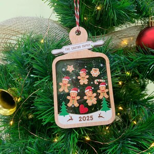 May include: Wooden Christmas ornament with a rectangular frame, featuring gingerbread people, stars, and a heart. The text "THE CASTRO FAMILY" is above the frame, and "2025" is at the bottom. The ornament hangs from a red and white string.