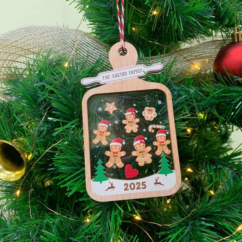 May include: Wooden Christmas ornament with a rectangular frame, featuring gingerbread people, stars, and a heart. The text "THE CASTRO FAMILY" is above the frame, and "2025" is at the bottom. The ornament hangs from a red and white string.