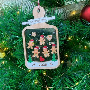 May include: Wooden Christmas ornament with a cutting board shape. It features gingerbread people, a star, and a heart. The text "THE CASTRO FAMILY" and "2025" are visible. The ornament hangs on a Christmas tree.