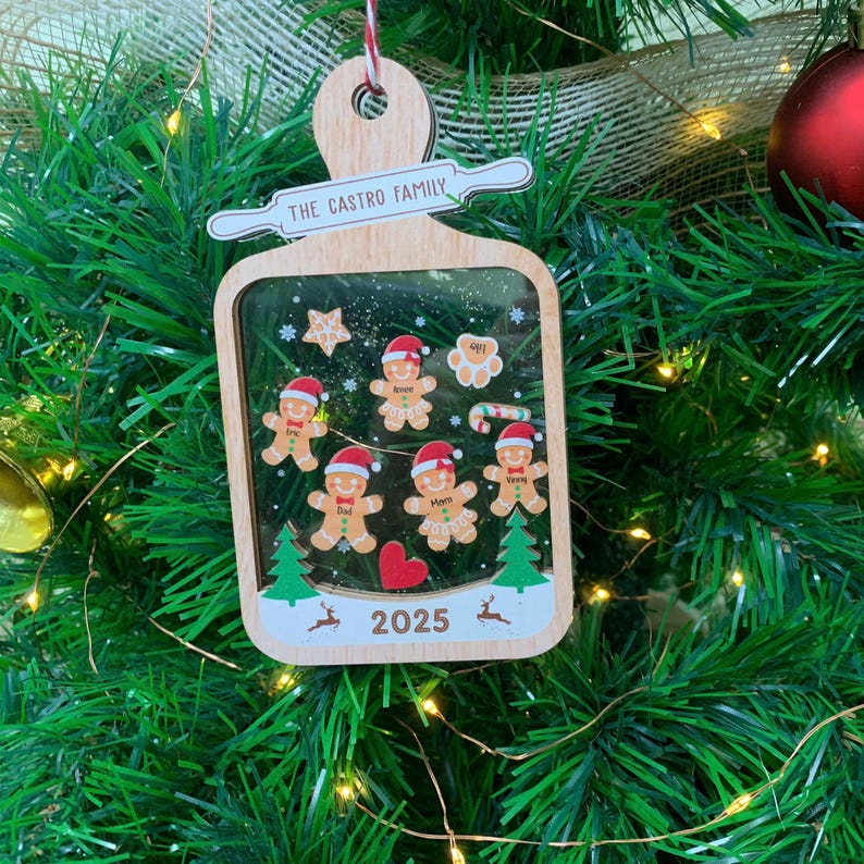May include: Wooden Christmas ornament with a cutting board shape. It features gingerbread people, a star, and a heart. The text "THE CASTRO FAMILY" and "2025" are visible. The ornament hangs on a Christmas tree.