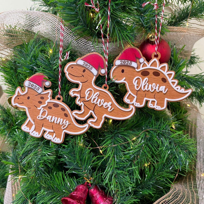 Op de afbeelding: Drie bruine dinosaurusornamenten met donkerbruine vlekken, elk met een rood-witte kerstmuts met het jaartal 2025 erop gedrukt. De ornamenten zijn gepersonaliseerd met de witte inscripties Danny, Oliver en Olivia en hangen aan rood-wit koord.