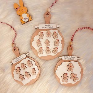 Puede incluir: Tres adornos de madera con forma de bandejas de galletas de jengibre con el texto "Nana's perfect batch", "Mama's perfect batch" y "Gigi & Poppop's perfect batch". Cada bandeja tiene figuras de pan de jengibre.
