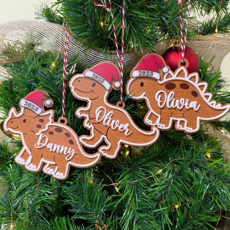 Op de afbeelding: Drie houten dinosaurusornamenten hangen in een kerstboom. Elke dinosaurus draagt een rode kerstmuts met het jaartal 2025 erop. De dinosaurussen zijn gepersonaliseerd met de witte in cursief geschreven namen Danny, Oliver en Olivia.