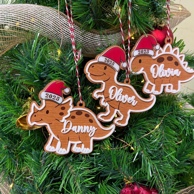 Op de afbeelding: Drie houten dinosaurusornamenten hangend aan rood-wit koord. Elke dinosaurus draagt een rode kerstmuts met het jaartal 2025 erop. De namen Danny, Oliver en Olivia staan in witte schrijfletters op de lichamen van de dinosaurussen afgebeeld.