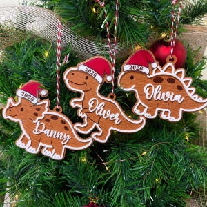 Op de afbeelding: Drie houten dinosaurusornamenten met een bruine houtnerfafwerking, elk met een rood-witte kerstmuts met het jaartal 2026 erop. De ornamenten zijn voorzien van de namen Danny, Oliver en Olivia in witte schrijfletters en hangen aan rood-wit koord.