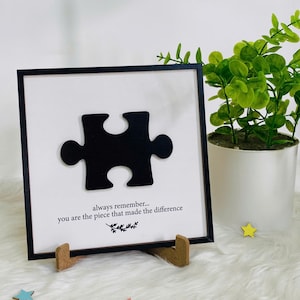 Pode incluir: Uma obra de arte emoldurada com uma peça de quebra-cabeça preta sobre um fundo branco. O texto diz "always remember... you are the piece that made the difference". A moldura preta contém a obra, que é exibida em um suporte de madeira.