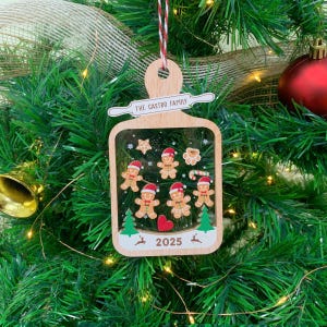 May include: Wooden Christmas ornament with a rectangular frame, featuring gingerbread people, stars, and a heart. The ornament has the text "THE CASTRO FAMILY" and "2025". It hangs from a red and white string.