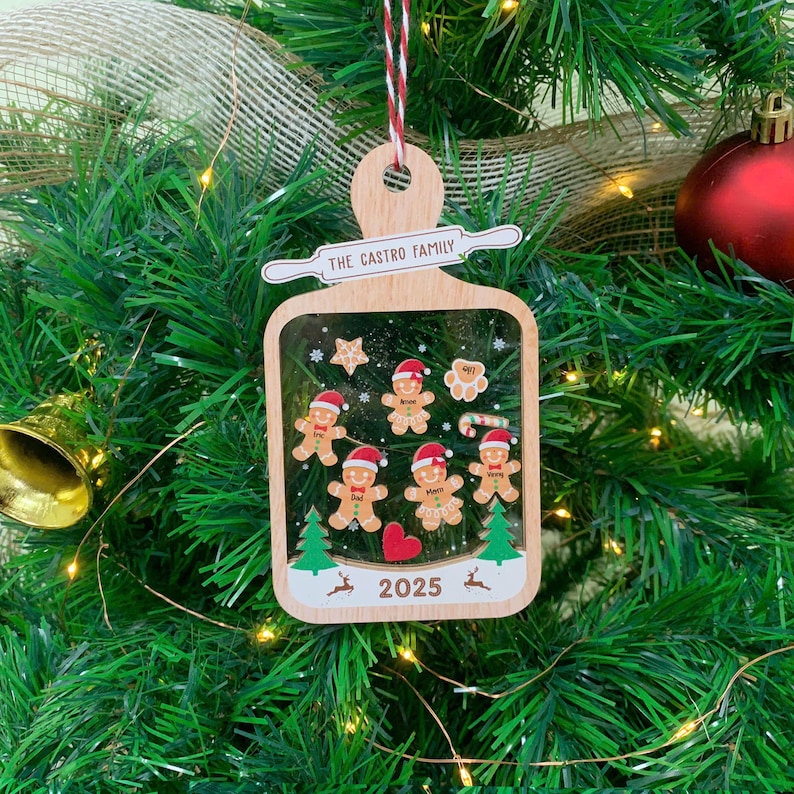 May include: Wooden Christmas ornament with a rectangular frame, featuring gingerbread people, stars, and a heart. The ornament has the text "THE CASTRO FAMILY" and "2025". It hangs from a red and white string.