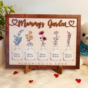 Peut inclure: Une pancarte en bois encadrée avec le texte "Mommy's Garden" et cinq illustrations florales dans des bouteilles en verre. Chaque bouteille porte un nom et une date imprimés. La pancarte est sur une surface blanche avec des cœurs rouges.