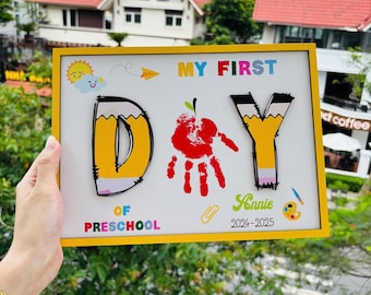 May include: A yellow-framed sign with the words "MY FIRST DAY OF PRESCHOOL" in colorful letters. The letter "D" is designed as a pencil, and a red handprint is in the center. The sign includes the name "Annie" and the year 2024-2025.