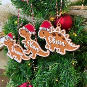 Op de afbeelding: Drie houten dinosaurusornamenten hangen in een kerstboom. Elke dinosaurus draagt een rood-witte kerstmuts met het jaartal 2025 erop gedrukt. De ornamenten zijn gepersonaliseerd met de namen Danny, Oliver en Olivia in een wit handschrift.