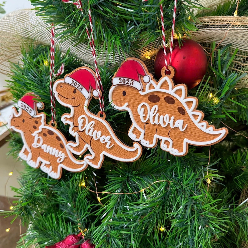 Op de afbeelding: Drie houten dinosaurusornamenten hangen in een kerstboom. Elke dinosaurus draagt een rood-witte kerstmuts met het jaartal 2025 erop gedrukt. De ornamenten zijn gepersonaliseerd met de namen Danny, Oliver en Olivia in een wit handschrift.