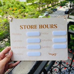 May include: A wooden store hours sign with the words "STORE HOURS" in brown lettering. The days of the week are listed below, with blank white rectangles for indicating the hours. The sign is held by a hand.