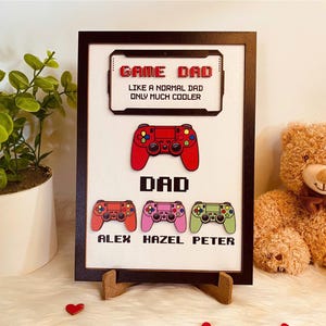 May include: A framed picture with a black frame and a white background. The picture says "Game Dad Like a normal dad only much cooler" and has a picture of a video game controller. Below the text are three more video game controllers with the names "Alex", "Hazel", and "Peter" underneath them.