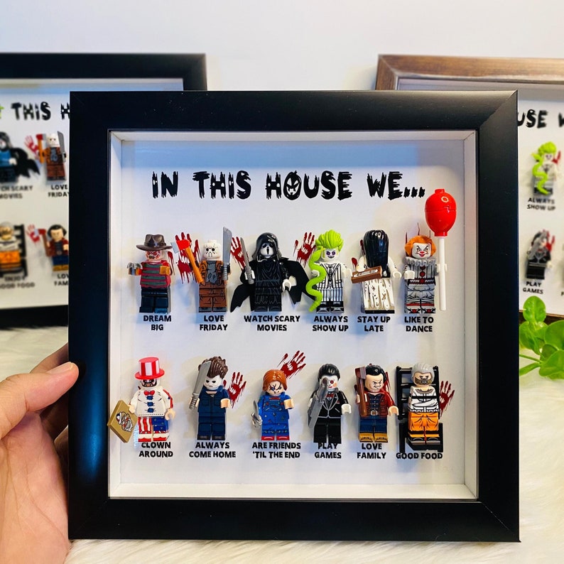 May include: A black framed shadow box with a white background. The shadow box contains a collection of 10 horror movie character Lego minifigures. The minifigures are arranged in two rows. The top row includes Freddy Krueger, Jason Voorhees, Ghostface, Michael Myers, and Pennywise. The bottom row includes Chucky, Leatherface, Samara Morgan, Hannibal Lecter, and Norman Bates. Each minifigure has a caption below it. The captions include: Dream Big, Love Friday, Watch Scary Movies, Always Show Up, Stay Up Late, Like to Dance, Clown Around, Always Come Home, Are Friends 'Til the End, Play Games, Love Family, Good Food.