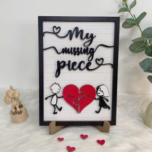May include: Black framed wooden sign with a white background and black lettering that reads "My missing piece". Two stick figures are holding a red heart shaped puzzle piece with the names "Tristan" and "Neil" written on them.