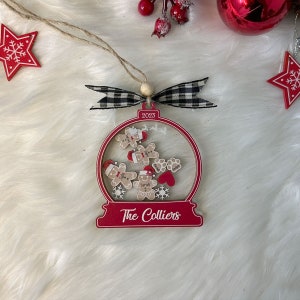 May include: A red and white snow globe ornament featuring the text 2023 and The Colliers. Inside are wooden gingerbread figures with red hats, paw prints, and a red heart. The ornament is topped with a black and white checkered ribbon and twine.