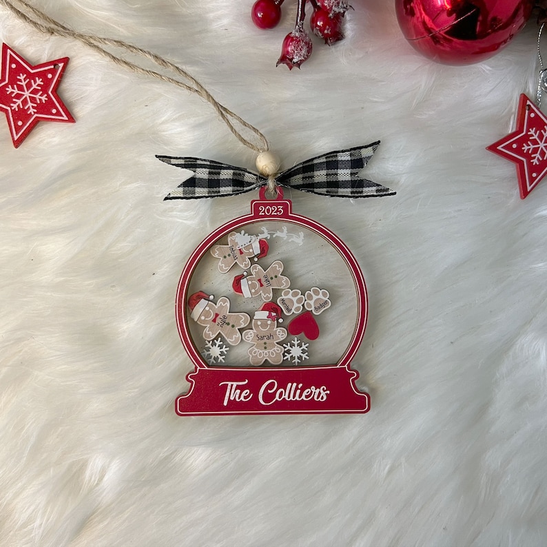 May include: A red and white snow globe ornament featuring the text 2023 and The Colliers. Inside are wooden gingerbread figures with red hats, paw prints, and a red heart. The ornament is topped with a black and white checkered ribbon and twine.