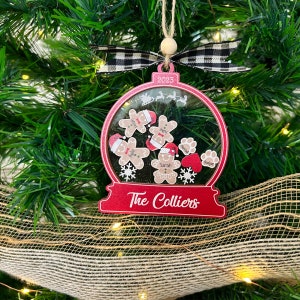 May include: A red, globe-shaped Christmas ornament featuring a clear center with gingerbread person cutouts, paw prints, and snowflakes. The ornament displays the text 2023 and The Colliers, topped with a black and white gingham ribbon bow.