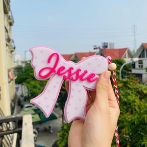 May include: A pink bow-shaped decoration with the name "Jessie" in bright pink script. The bow has a white base with small pink hearts and a pink outline. A red and white string is attached.
