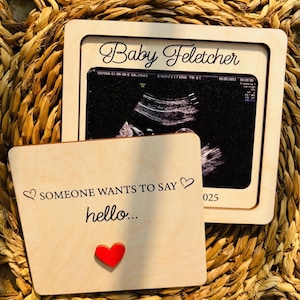 Könnte beinhalten: Ein Holzrahmen für Ultraschallbilder mit dem Text "Baby Feletcher" und einem Ultraschallbild. Eine kleinere Holzkarte lautet "SOMEONE WANTS TO SAY hello..." mit einem roten Herzen. Die Artikel befinden sich auf einem gewebten Hintergrund.