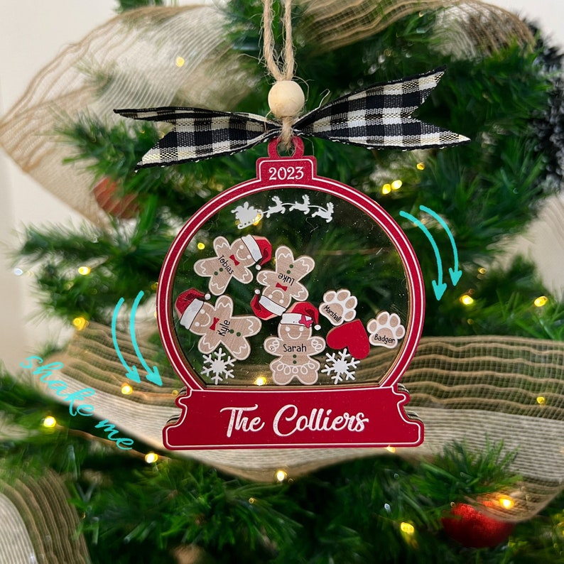 May include: A red and clear snow globe ornament featuring the text 2023 and The Colliers. Inside are gingerbread figures with names Tobias, Luke, Kyle, and Sarah, plus paw prints labeled Marshall and Badger, a red heart, and snowflakes.