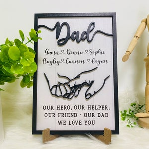 May include: Black framed wooden sign with white background. The sign features a black silhouette of two hands holding each other. The text "Dad" is in a large, stylized font at the top of the sign. Below the hands, the text reads "Our hero, our helper, our friend - our dad. We love you."  The names "Gavin, Donna, Sophie, Hayley, Cannon, and Logan" are listed below the word "Dad".