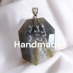 Peut inclure: Un pendentif en agate gris avec une bélière en argent. L'agate a un motif blanc et est inscrite de texte blanc. Le pendentif est fait main.