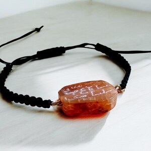 May include: A black cord bracelet with a brown, rectangular stone featuring white inscription. The stone is attached to the cord with a gold wire.