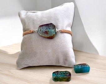 May include: A tan braided bracelet with a large, green, brown, and blue stone pendant. The stone has white markings on it. There are two smaller, green, brown, and blue stones with white markings on them, sitting on a wooden surface.