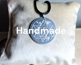 May include: A silver pendant necklace with a round, engraved charm featuring a six-pointed star and Hebrew text. The pendant is on a black cord and is displayed on a light gray velvet pillow.