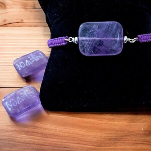 May include: A purple amethyst gemstone bracelet with a braided cord and silver accents. The bracelet is displayed on a black velvet cushion.