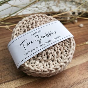 Puede incluir: Un exfoliante facial hecho a mano, de color beige y tejido a ganchillo, con una etiqueta blanca envuelta. La etiqueta dice "Face Scrubbies" y "100% algodón - ecológico - reutilizable". El exfoliante es circular y está hecho de un hilo grueso y texturizado.