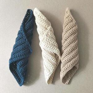 May include: Three handmade crochet spiral-shaped washcloths in shades of blue, cream, and beige. The washcloths are arranged on a light gray surface, showcasing their textured, woven design. The soft, absorbent cloths are ideal for kitchen or bathroom use.