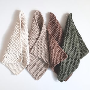 May include: Four knitted kitchen towels in varying neutral colors: cream, tan, brown, and olive green. The towels are hanging against a white background. Each towel has a textured, woven appearance, and is folded into a triangle shape.