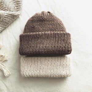 May include: Two knitted beanies, one brown and one beige, are stacked on top of each other. The brown beanie is on top, with a folded brim. A herringbone patterned scarf and a tassel are in the background.