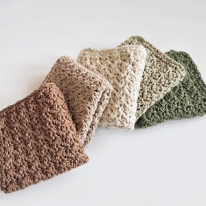 May include: A collection of five crocheted washcloths in varying neutral colors: brown, beige, cream, olive green, and dark green. The washcloths are folded and arranged in a diagonal line, showcasing their textured, handmade appearance.