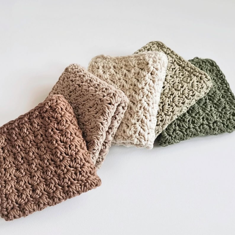 May include: A collection of five crocheted washcloths in varying neutral colors: brown, beige, cream, olive green, and dark green. The washcloths are folded and arranged in a diagonal line, showcasing their textured, handmade appearance.