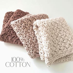 May include: Three folded, crocheted washcloths in shades of brown and cream. The washcloths are made of 100% cotton and have a textured, patterned design. The text "100% COTTON" is printed in a light gray color.