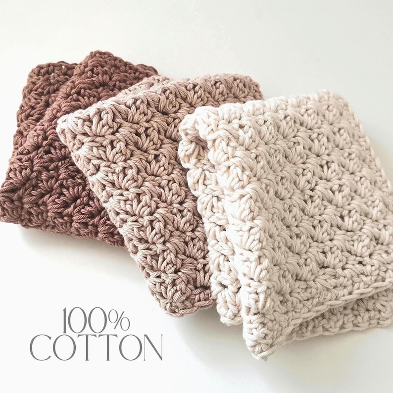 May include: Three folded, crocheted washcloths in shades of brown and cream. The washcloths are made of 100% cotton and have a textured, patterned design. The text "100% COTTON" is printed in a light gray color.