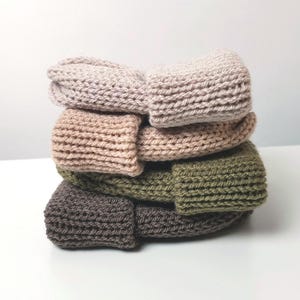 May include: A pile of six knitted beanies in shades of beige, tan, olive green, and brown. The beanies are folded over, displaying the ribbed knit texture. The hats are stacked on top of each other.