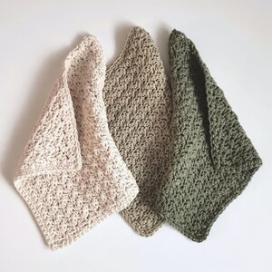 May include: Three handmade crochet washcloths in varying neutral tones: light beige, tan, and olive green. Each washcloth is folded, showcasing the textured stitch pattern. The soft, natural colors and handmade quality suggest a focus on eco-friendly and home decor items.