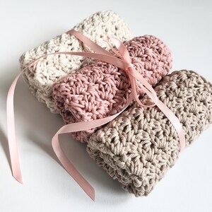 May include: A stack of three crocheted washcloths in cream, dusty rose, and taupe, tied with a pale pink satin ribbon. The washcloths are made of a textured, looped yarn, and are neatly folded. The overall aesthetic is soft and natural.